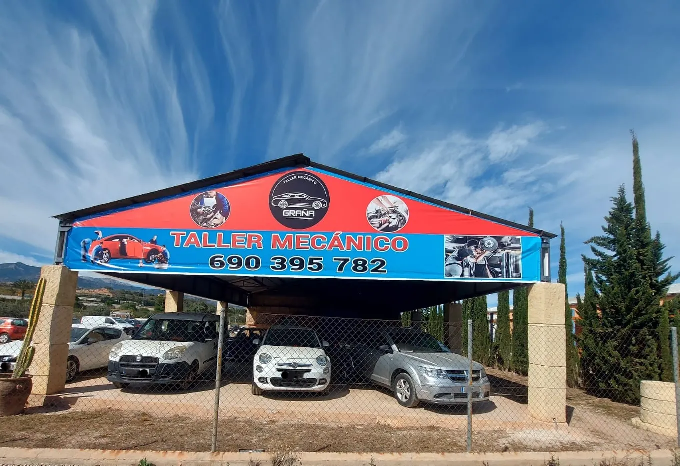El taller visto desde la carretera, con la lona de Taller Mecánico Graña sobre el porche y varios coches aparcados.