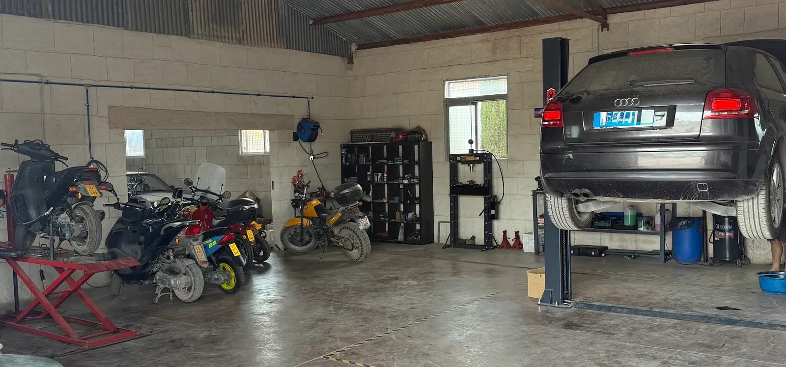 Interior del taller: una fila de motos y scooters a la izquierda y un coche subido al elevador de dos columnas a la derecha.